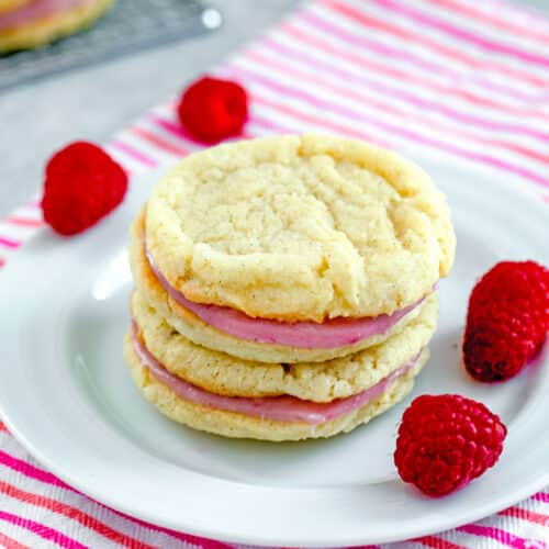 Closeup of a stack of sugar cookies with raspberry cream filling with fresh raspberries all around.