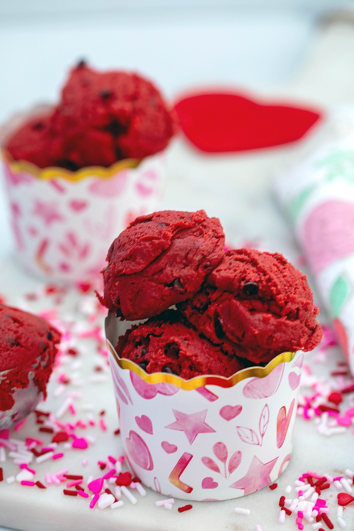 Head-on view of red velvet cookie dough in pink and red heart cup with sprinkles all around.