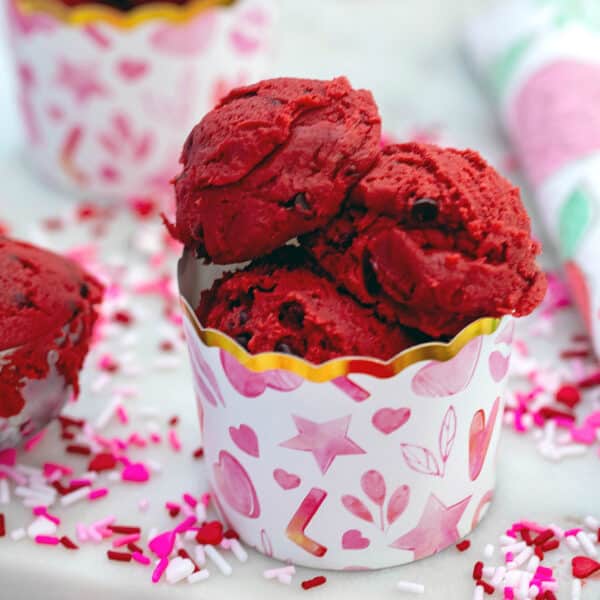 Closeup view of red velvet cookie dough in a small paper cup with red and pink sprinkles all around.