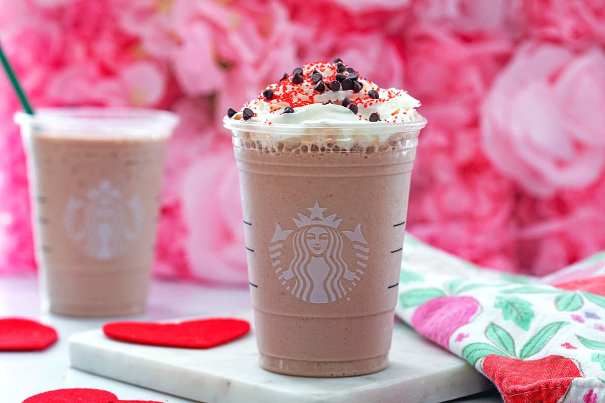 Landscape head-on view of a Red Velvet Frappuccino with a second in the background and red felt hearts all around.
