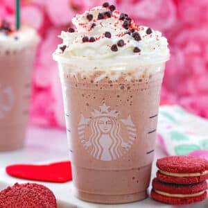Closeup view of a red velvet Frappuccino topped with whipped cream and chocolate chips with red velvet cookies on the side.