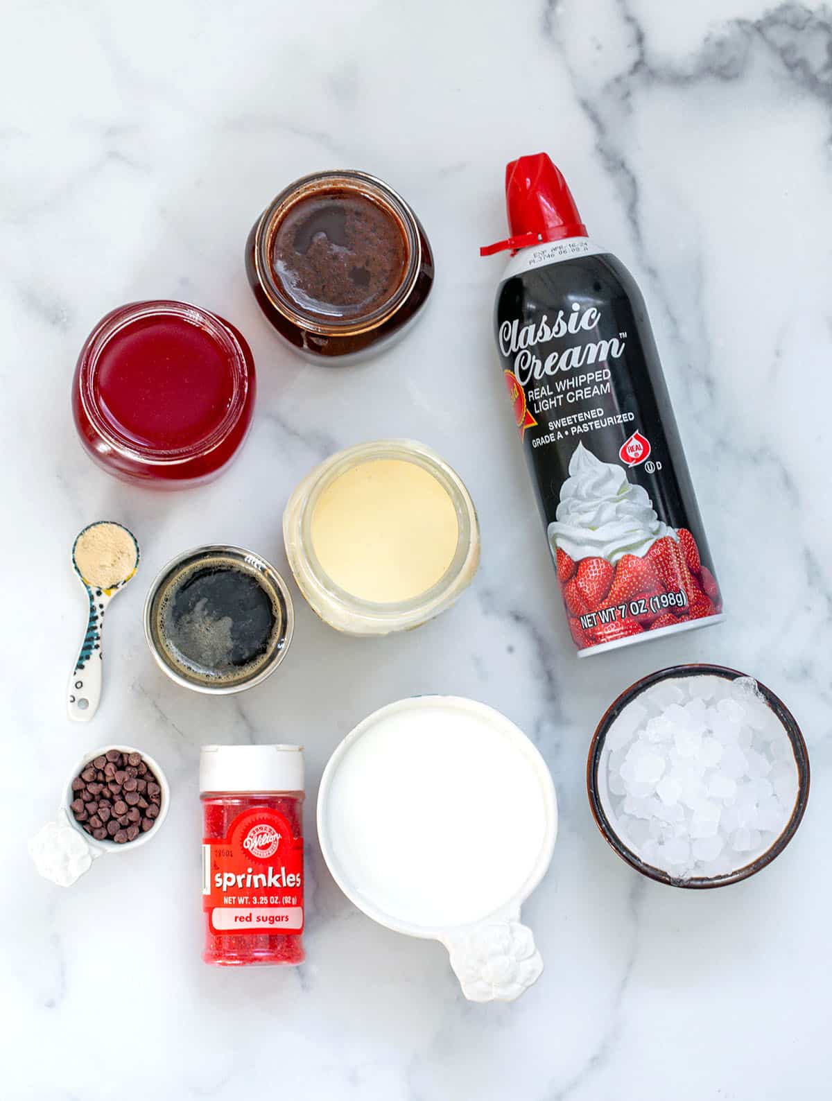 Ingredients on marble surface, including whipped cream, milk, ice, white mocha sauce, chocolate mocha sauce, raspberry syrup, espresso, chocolate chips, xanthan gum and red sprinkles.