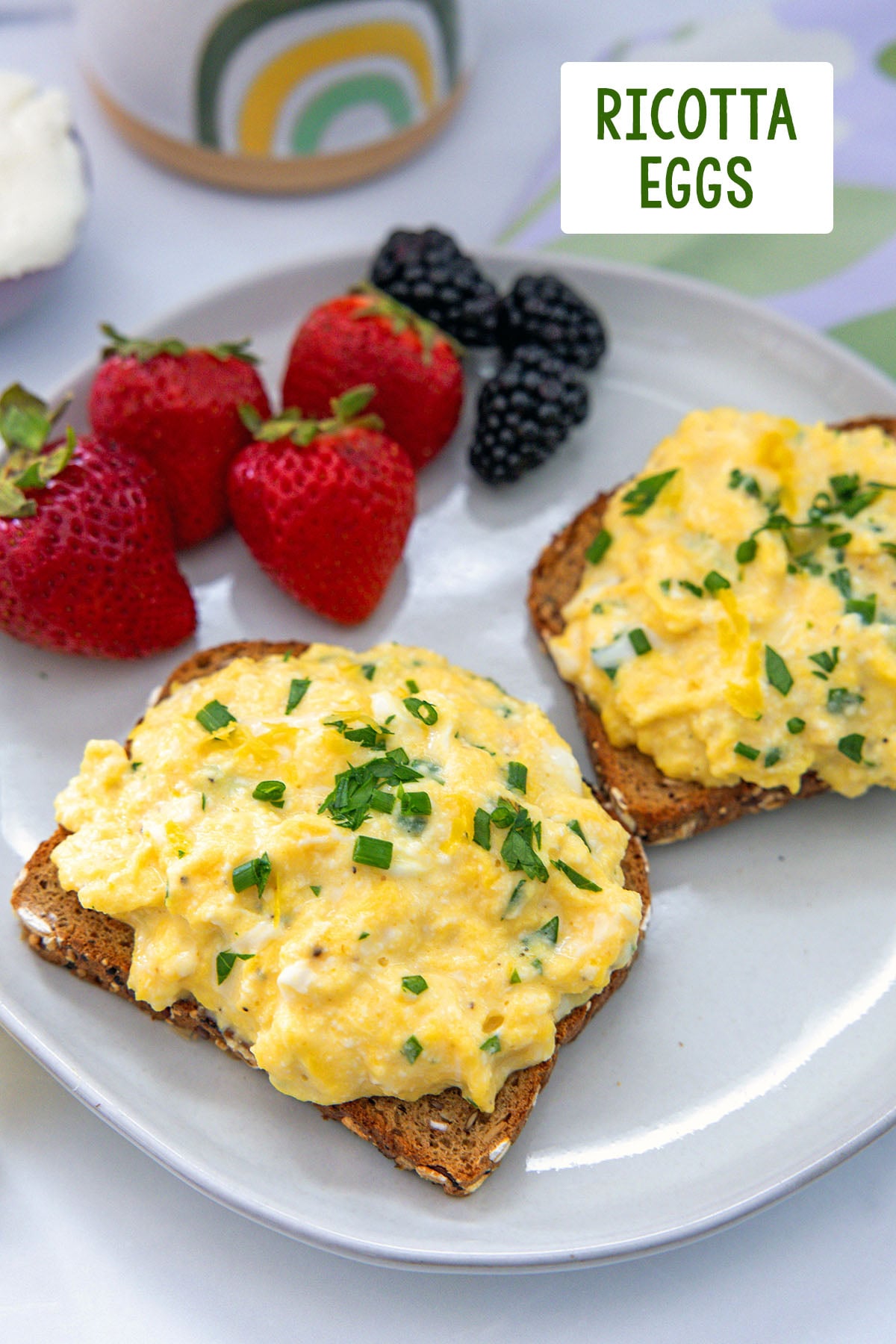 Two slices of toast topped with ricotta eggs with berries in background and recipe title at top.