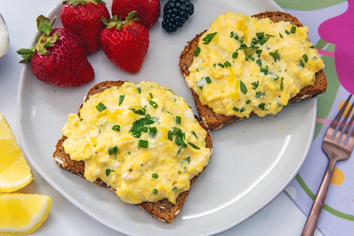 Landscape overhead view of a plate with two slices of toast topped with ricotta scrambled eggs with sliced lemons, strawberries, and blackberries in background.