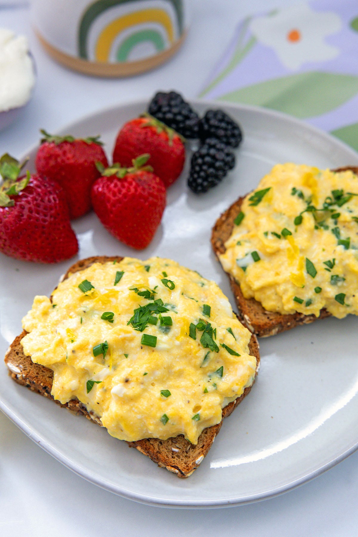 Two slices of toast topped with ricotta scrambled eggs with berries in background.