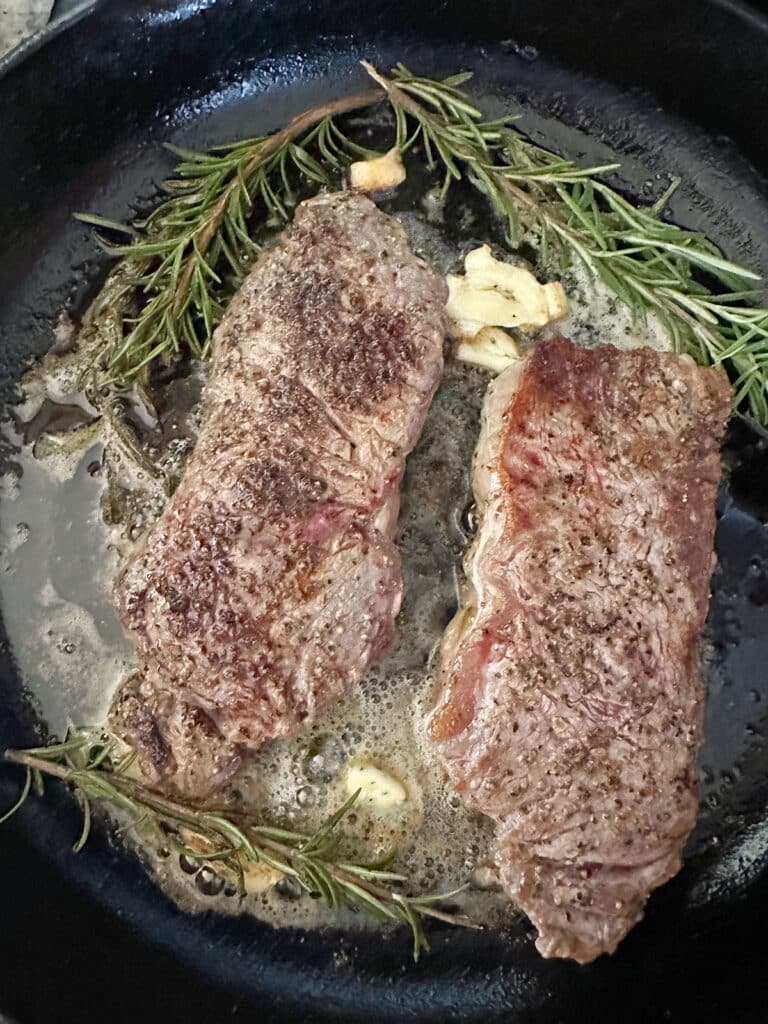 Two steaks cooking in skillet with sprigs of rosemary, crushed garlic, and buttter.