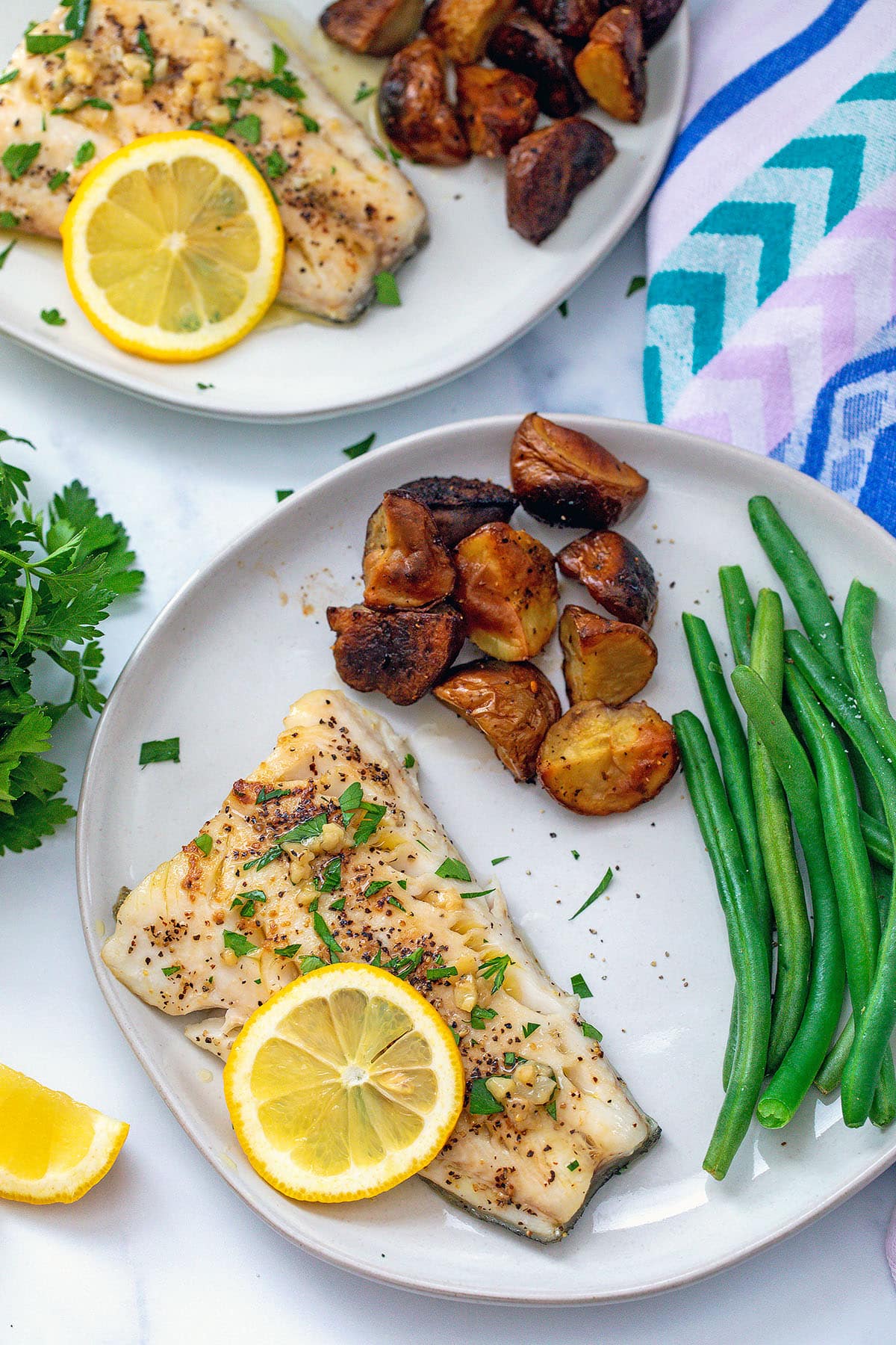 Sablefish with garlic butter on plate with green beans and potatoes with second plate in background.