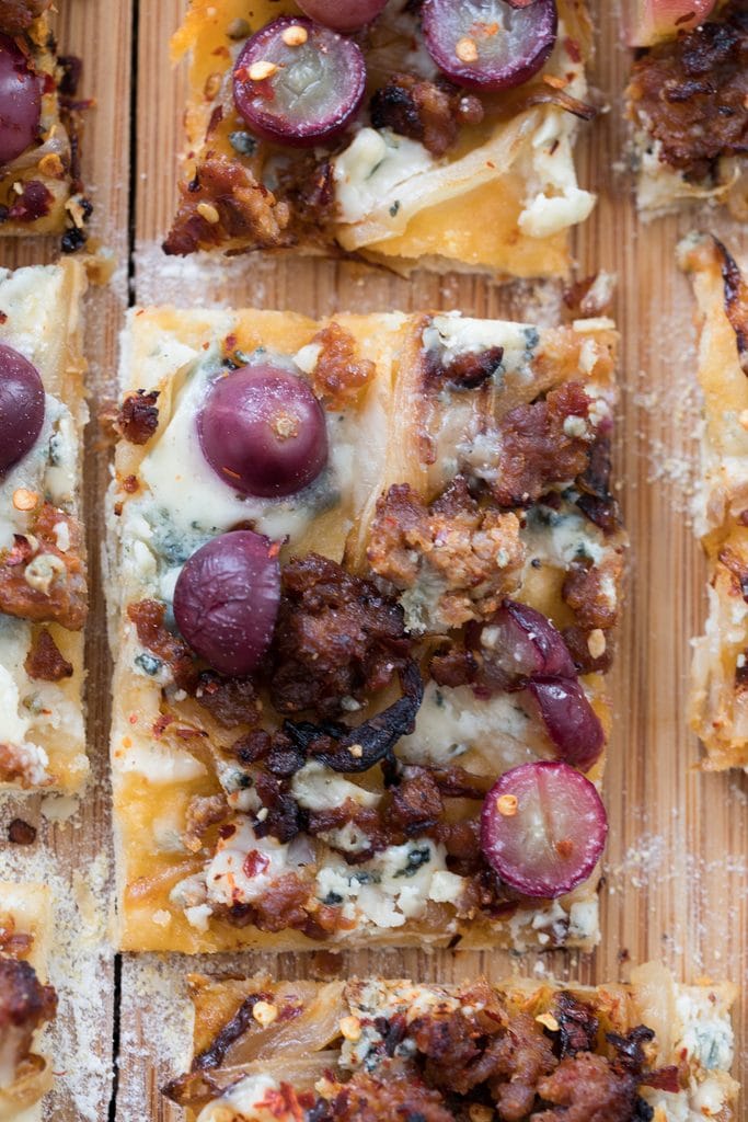 A close-up shot from above of a square slice of sausage, grape, and gorgonzola flatbread pizza