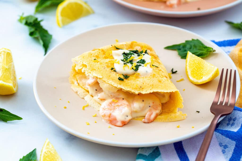 Landscape head-on view of a crepe filled with creamy shrimp and toped with cream sauce with lemon wedges and basil in background and fork on plate.