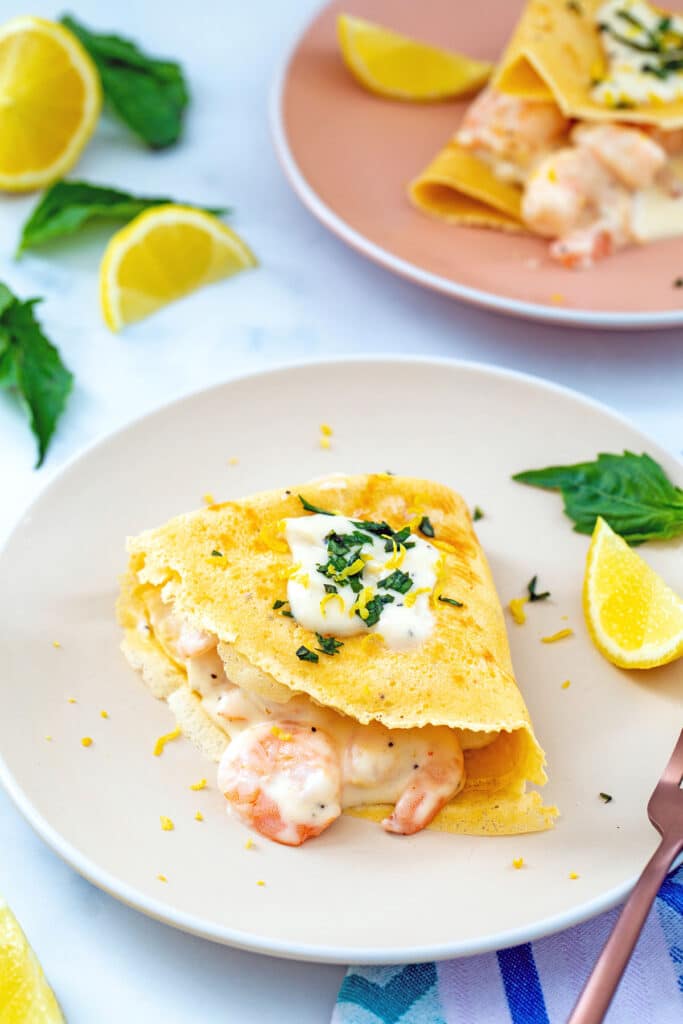 Crepe filled with creamy shrimp and topped with cream sauce and basil on a plate with second plate and lemon wedges in background.