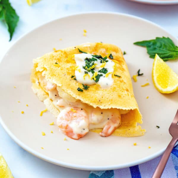 Closeup view of a shrimp crepe topped with cream sauce and basil on a plate with lemon wedge and basil.