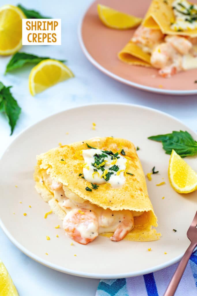 Crepe filled with creamy shrimp and topped with cream sauce and basil on a plate with second plate and lemon wedges in background and recipe title at top.