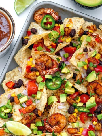 Shrimp nachos on a sheet pan with salsa, limes, and avocado in background.