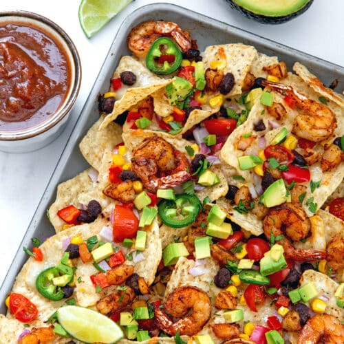 Shrimp nachos on a sheet pan with salsa, limes, and avocado in background.