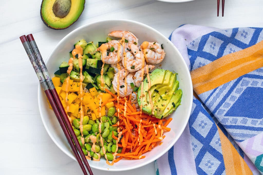 Landscape overhead view of a shrimp poke bowl with mango, avocado, edamame, carrots, mango, and cucumber with chopsticks on bowl.