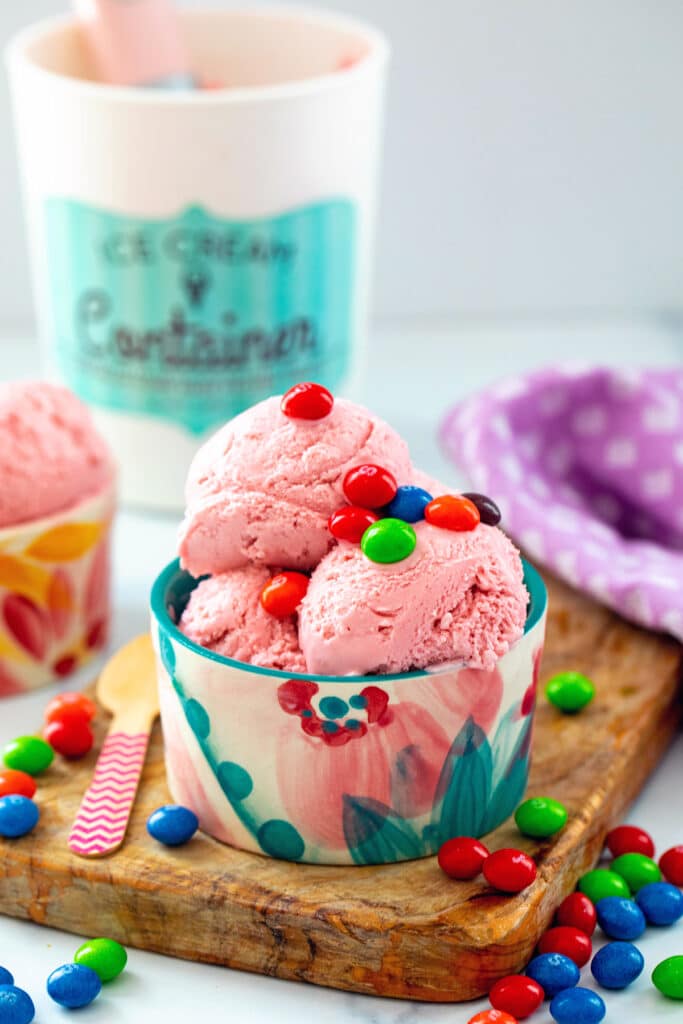 Small bowl of Skittles ice cream with candy all around and more ice cream in background.