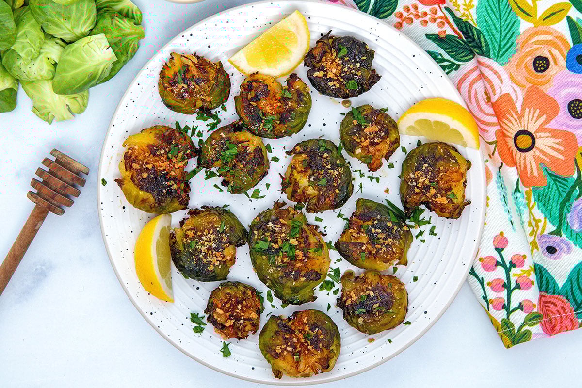 Overhead landscape view of smashed brussels sprouts on white plate with lemon wedges and honey stick and sprouts in background.