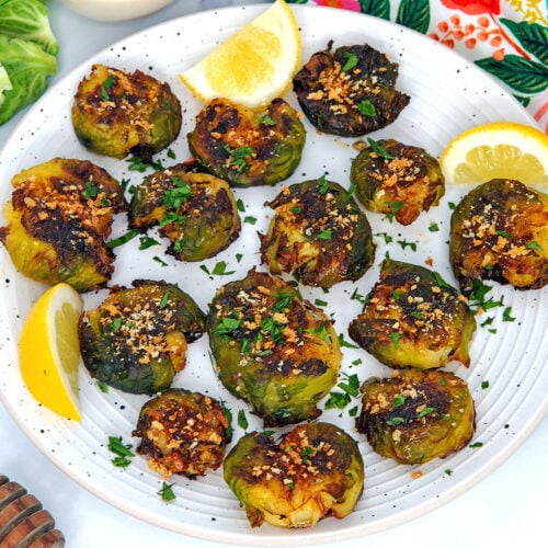Closeup view of smashed brussels sprouts on a white plate with lemon wedges.