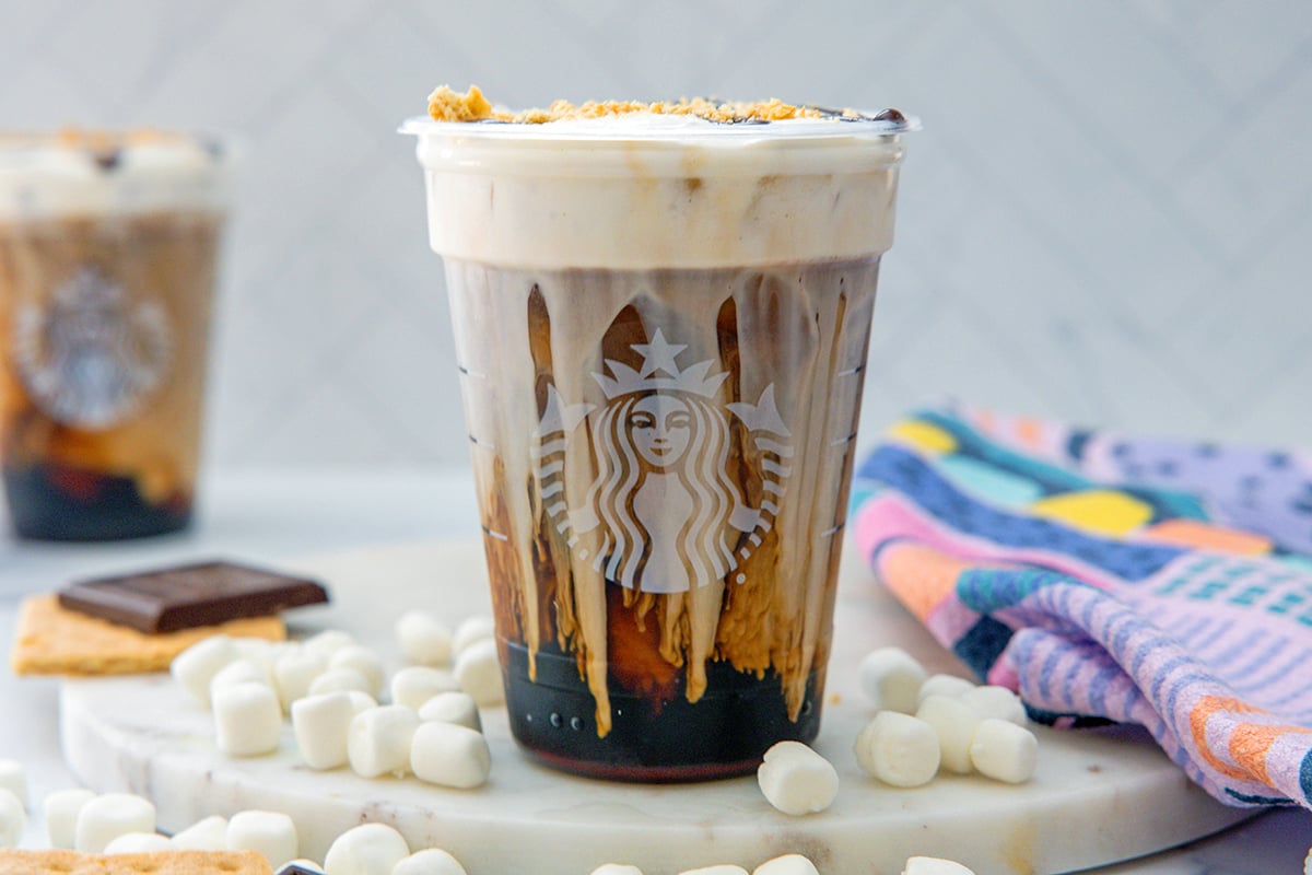 Landscape head-on view of a S'mores Cold Brew with second drink in background and mini marshmallows, graham crackers, and chocolate bars all around.
