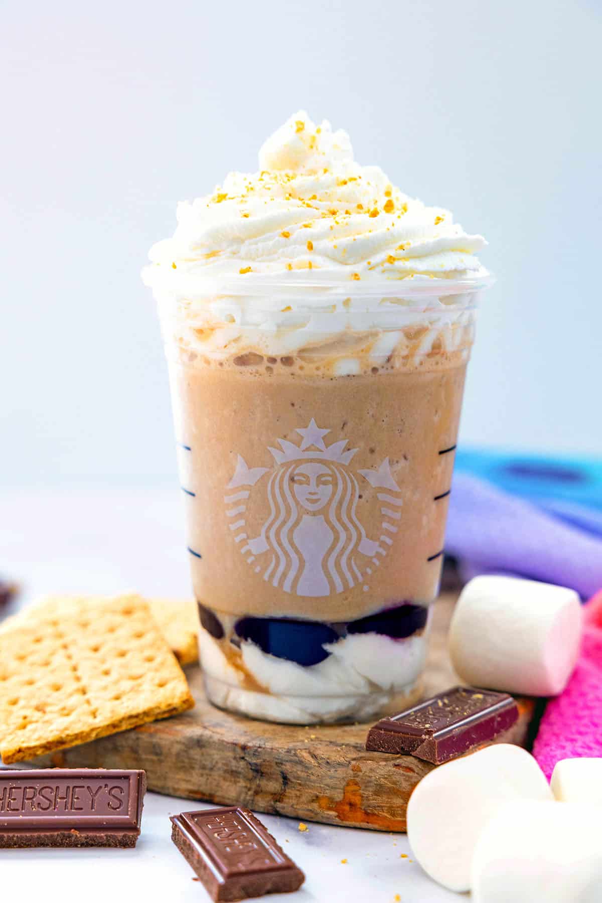 Head-on view of a S'mores Frappuccino in a Starbucks cup with chocolate bars, graham cracker squares, and marshmallows all around.
