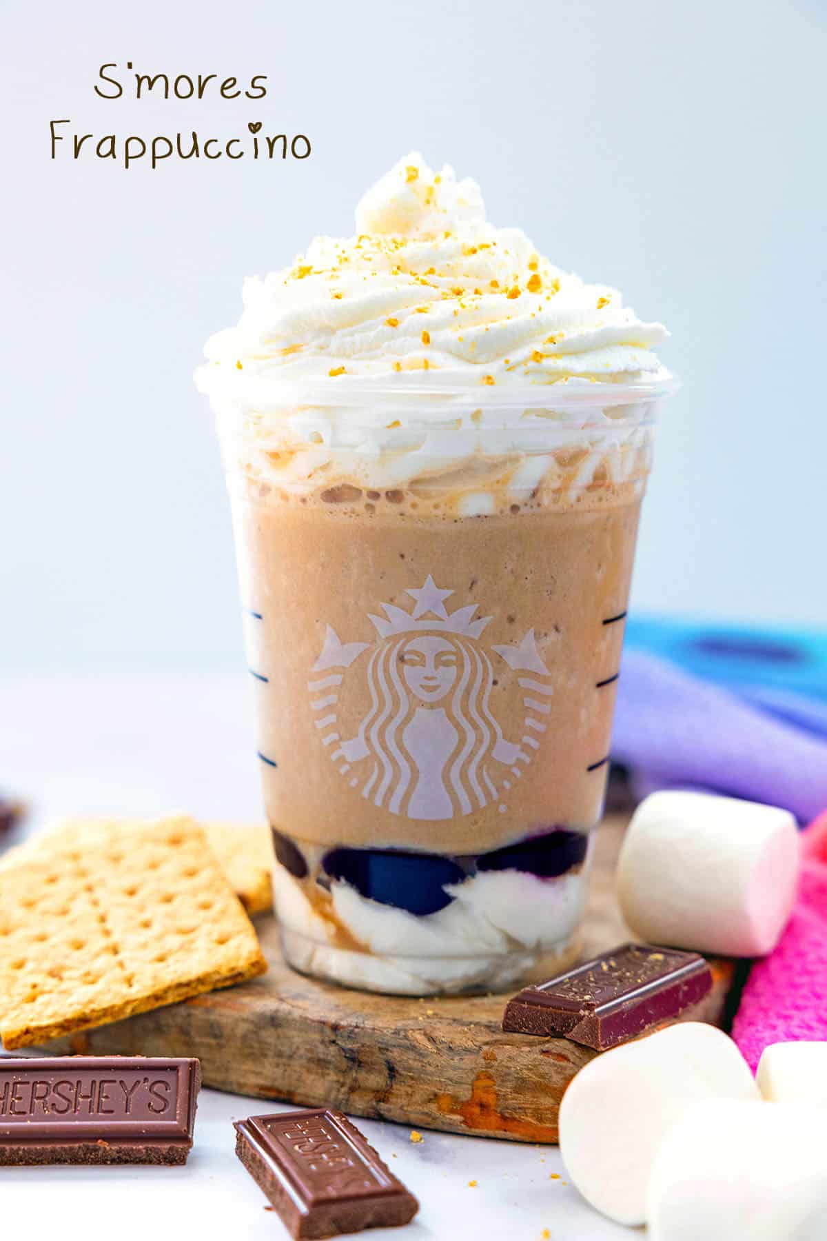 Head-on view of a s'mores Frappuccino in a Starbucks cup surrounded by graham crackers, chocolate bars, and marshmallows with recipe title at top.