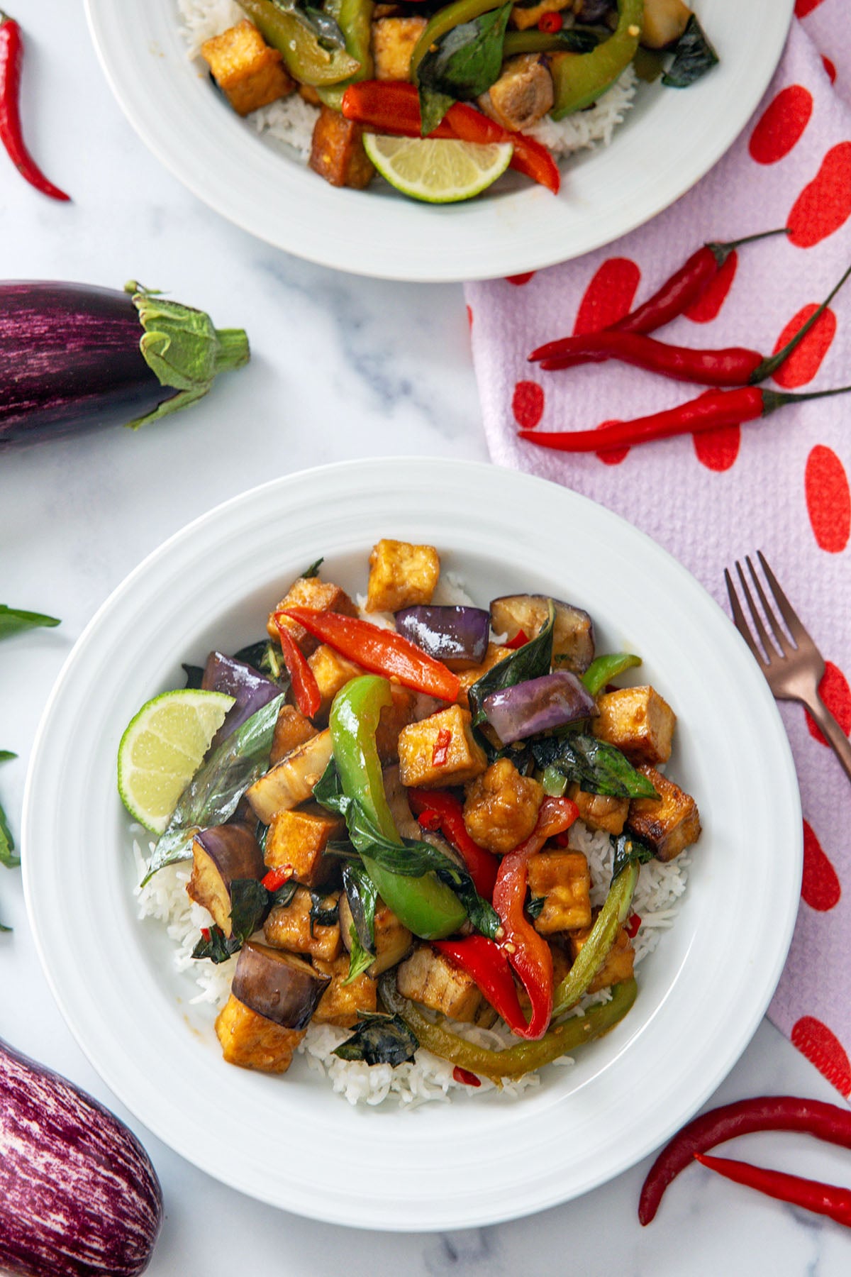 Spicy basil eggplant with tofu over rice on a white plate with peppers and mini eggplant in background.