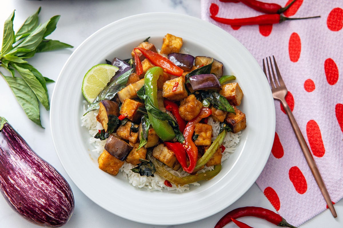 Landscape overhead view of spicy basil eggplant with tofu in white dish with Japanese eggplant, basil, hot peppers, and fork on the side.