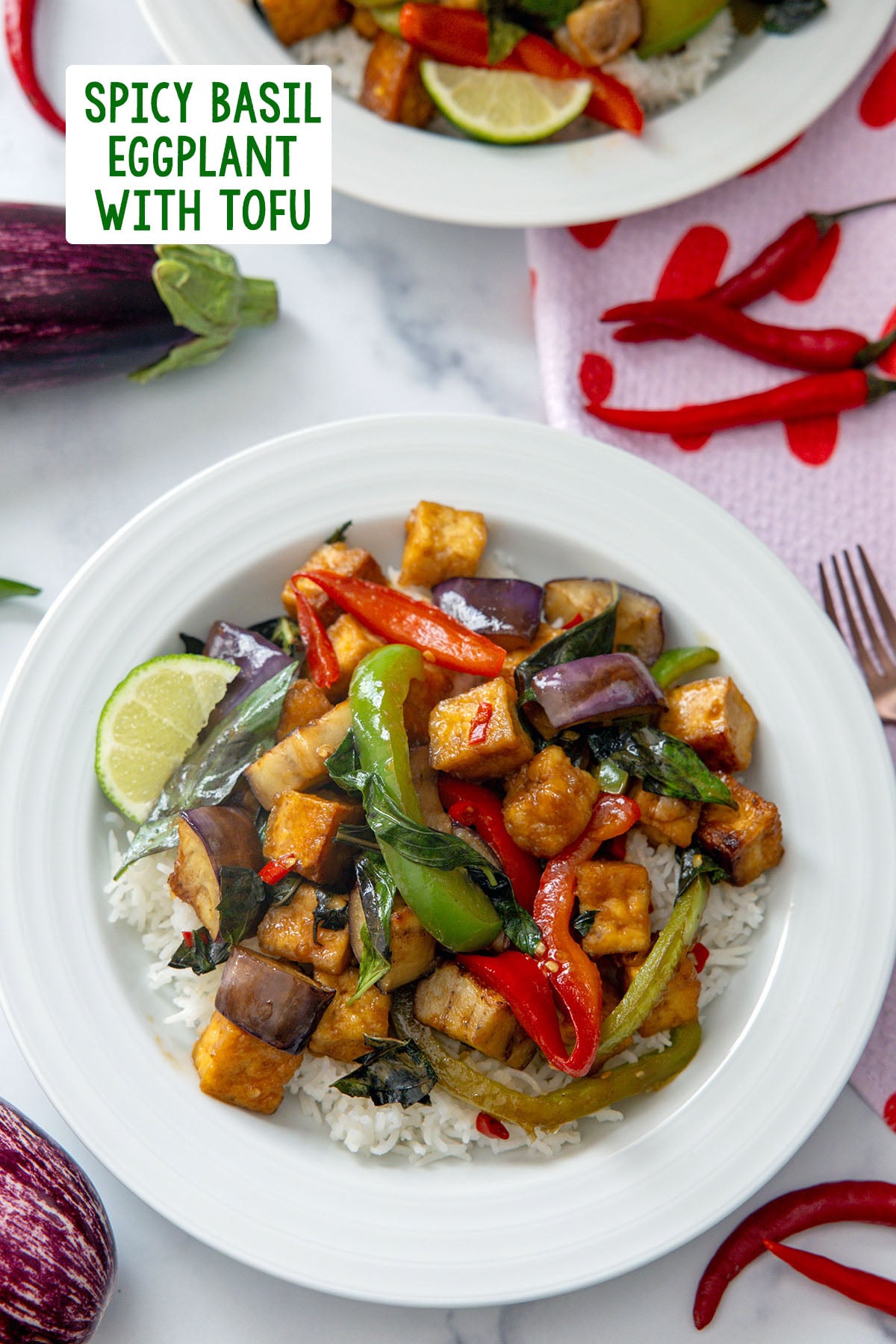 Spicy basil eggplant with tofu over rice on a white plate with peppers and mini eggplant in background and recipe title at top.