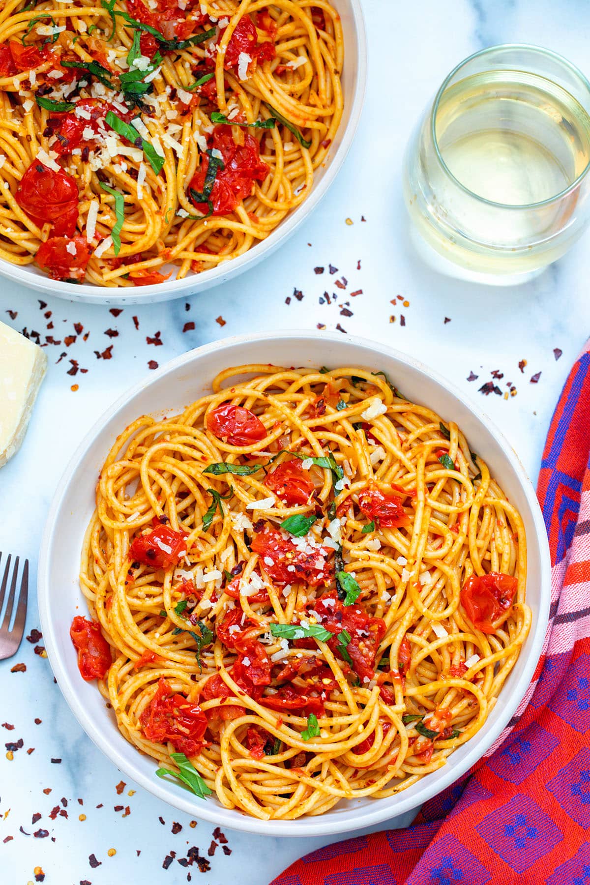 Two bowls of spicy spaghetti with fresh tomatoes and basil with red pepper flakes all around and a glass in the background.