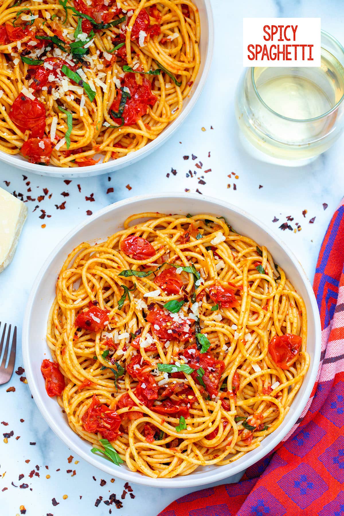 Two bowls of spicy spaghetti with fresh tomatoes and basil with red pepper flakes all around, a glass in the background and recipe title at top.