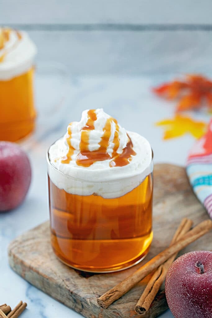 Caramel apple spice drink in a mug with whipped cream, caramel sauce, and cinnamon sticks and apples on the side.