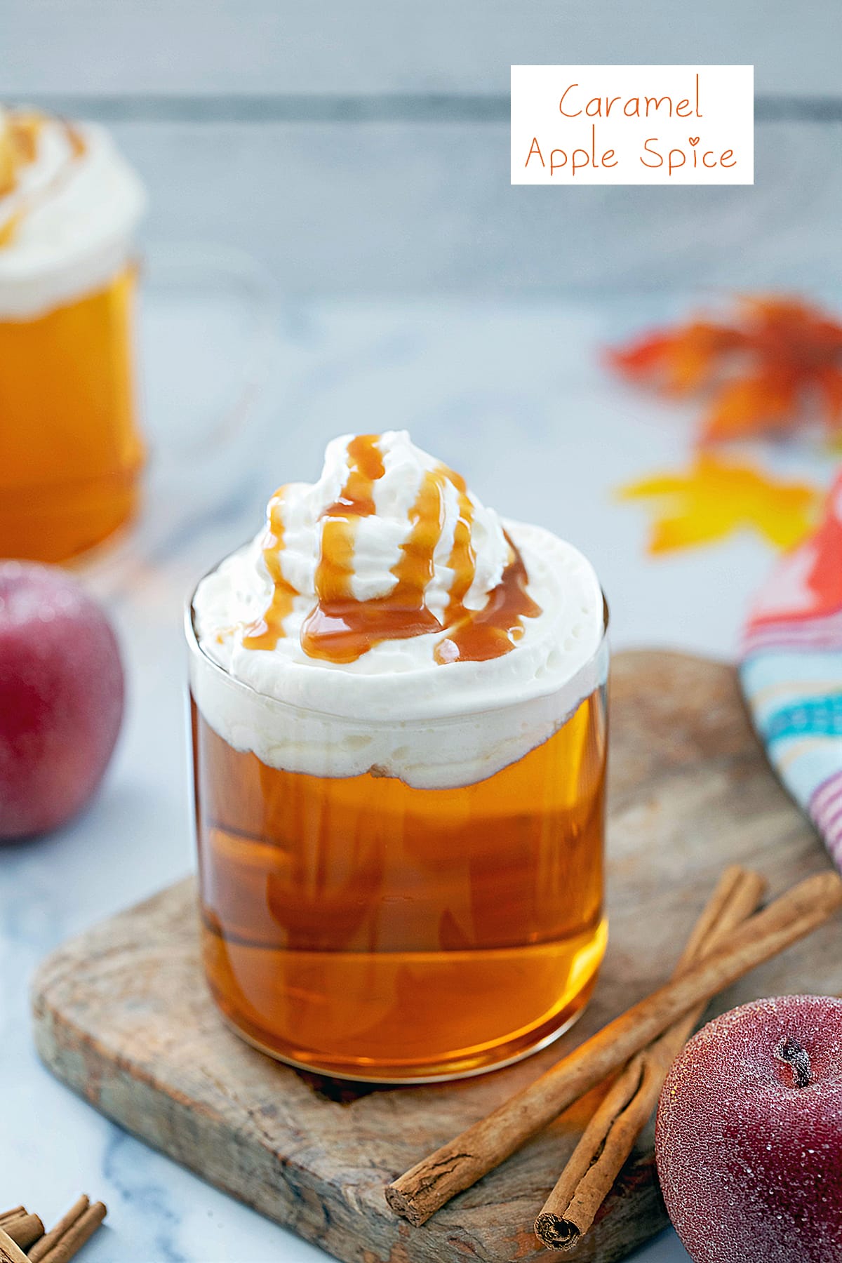 Caramel apple spice drink in a mug with whipped cream, caramel sauce, and cinnamon sticks and apples on the side with recipe title at top.
