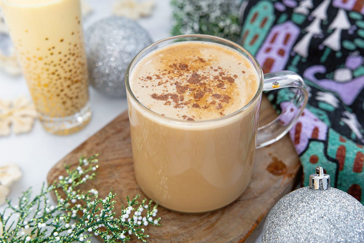 Overhead landscape view of an eggnog latte in a clear mug with glass of eggnog in background.