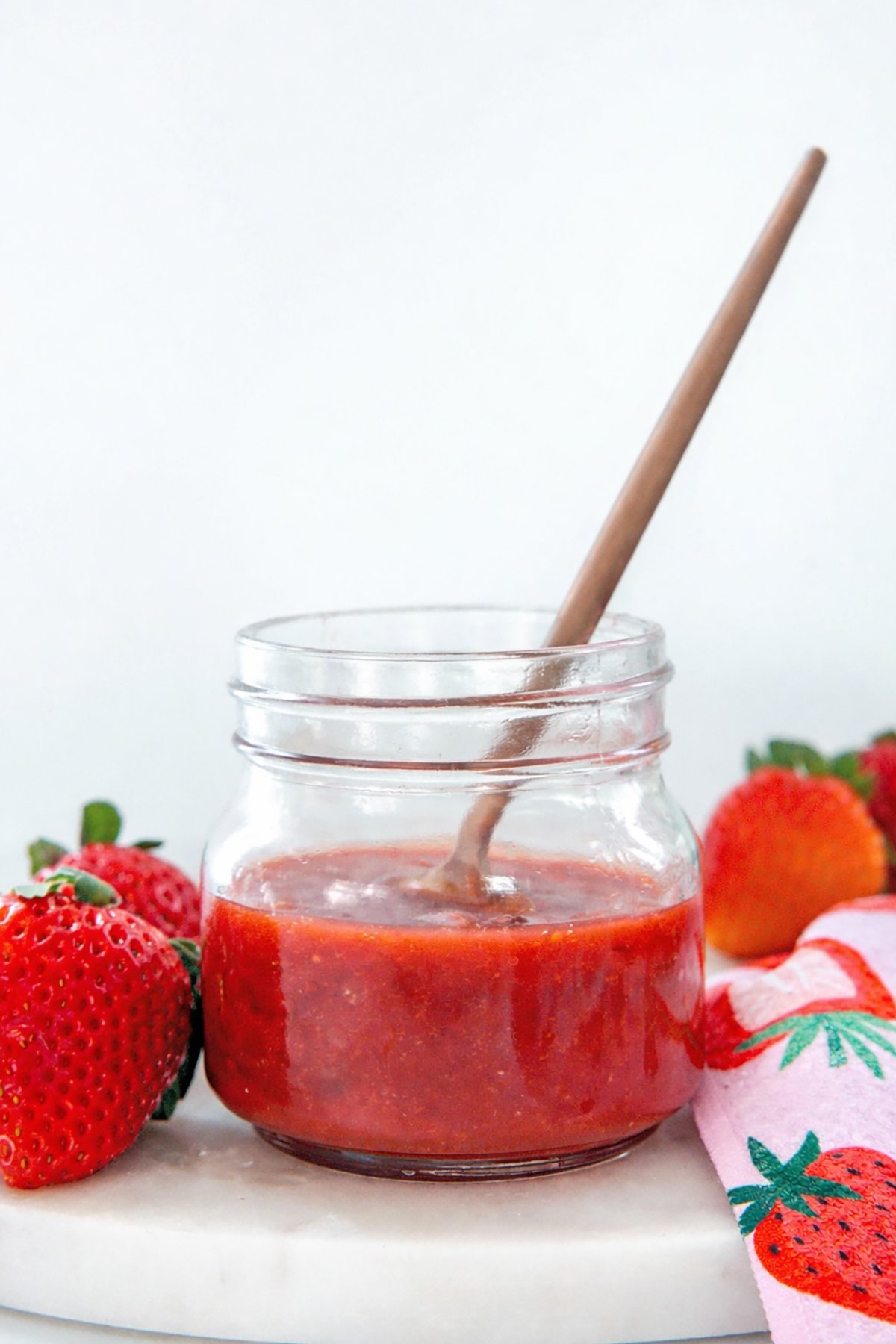 Head-on view of a small jar with homemade Starbucks strawberry puree in it and fresh strawberries all around.