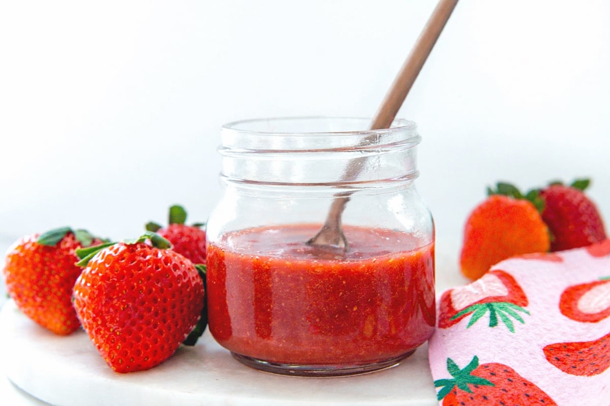 Head-on landscape view of a small jar of homemade Starbucks strawberry puree with fresh strawberries all around.