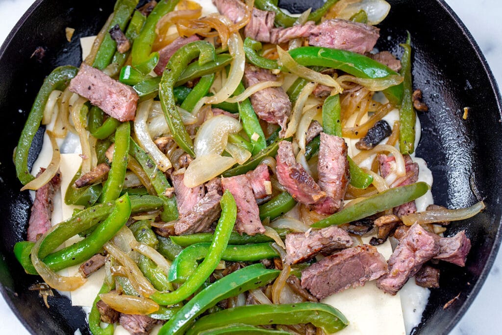 Steak, peppers, and onions over noodles in cast iron skillet.