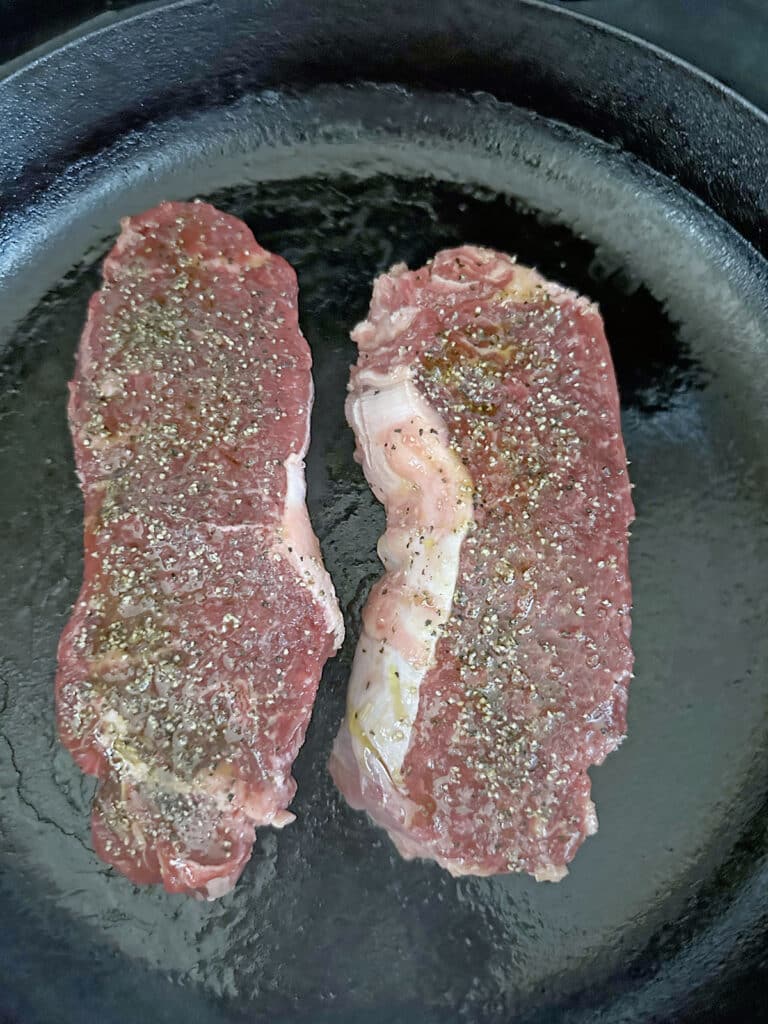 Two NY strip steaks cooking in cast iron skillet.