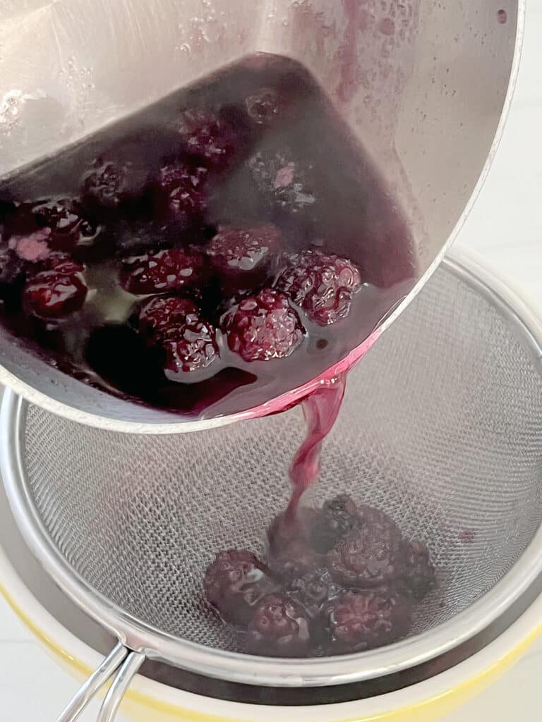 Blackberry syrup being poured from saucepan through strainer into a bowl.