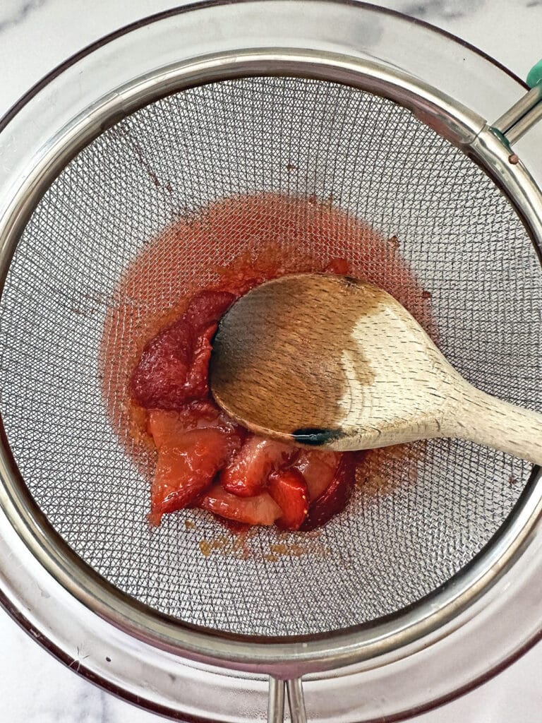 Wooden spoon pressing down on strawberries in strainer.