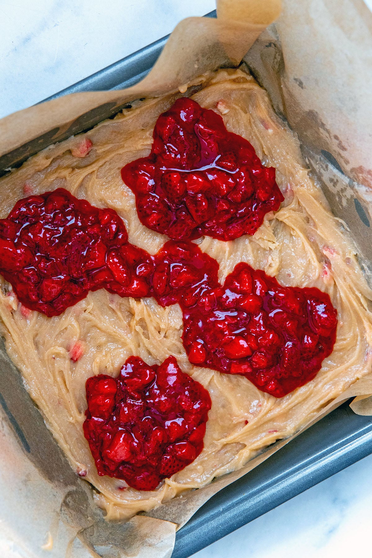 Strawberry brownie batter spread on the bottom of a pan with dollops of strawberry jam on top.
