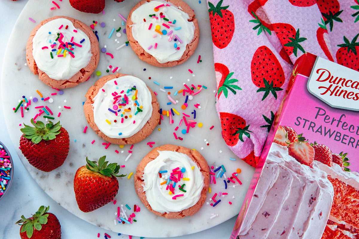 Landscape overhead view of frosted strawberry cookies with strawberries, sprinkles, and box of strawberry cake mix.