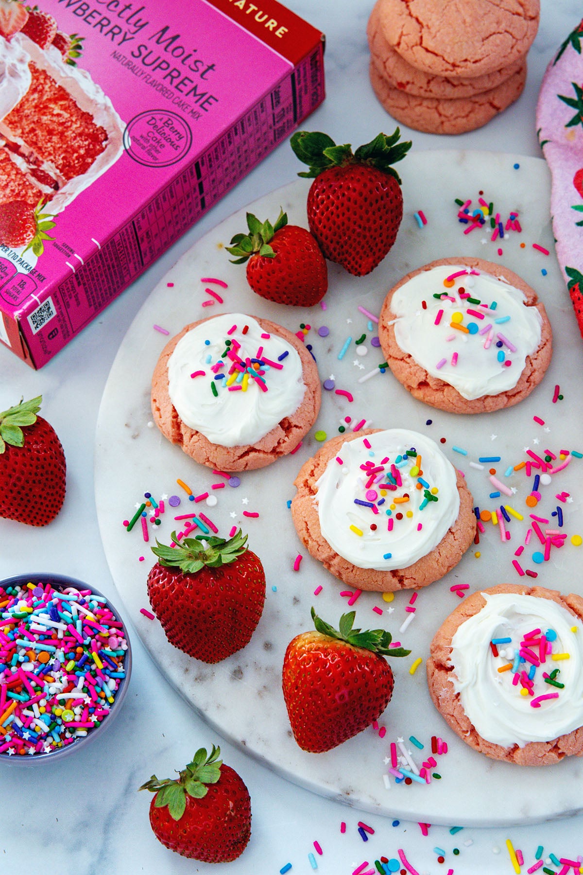 Overhead view of frosted strawberry cookies on marble tray with fresh strawberries and sprinkles all around and box of cake mix in the background.