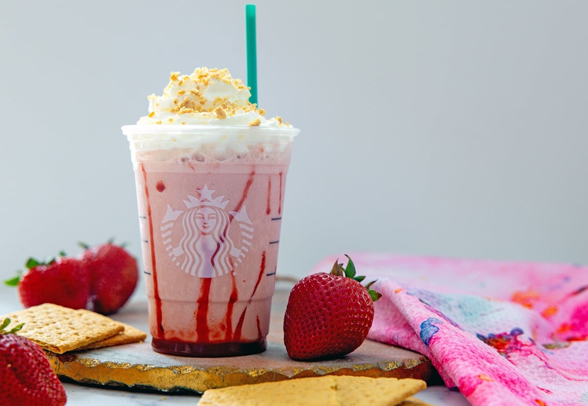 Landscape head-on view of a Strawberry Cheesecake Frappuccino on a wooden board with whole strawberries and graham crackers in background.