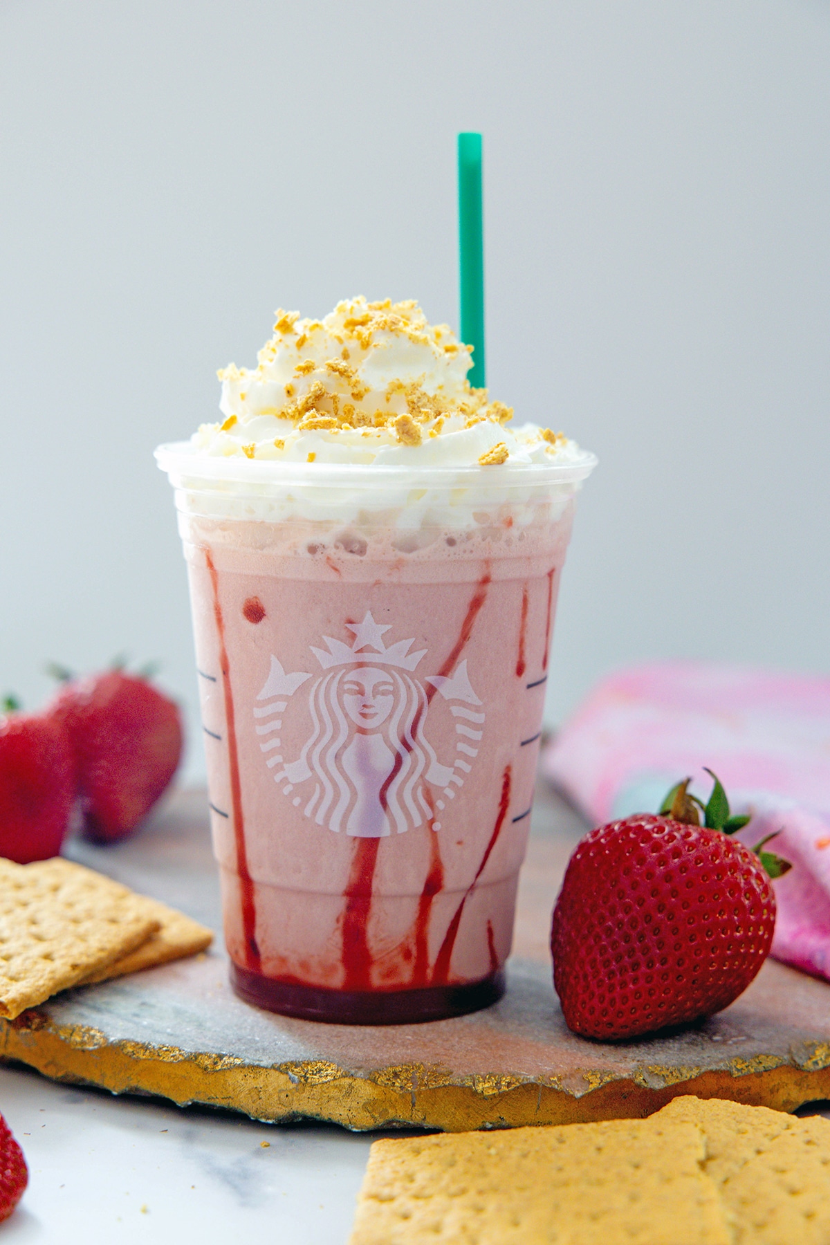 Head-on view of a Strawberry Cheesecake Frappuccino with a green straw with strawberries and graham crackers all around.