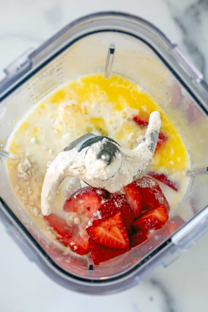 Overhead view of flour, sliced strawberries, eggs, and milk in blender.