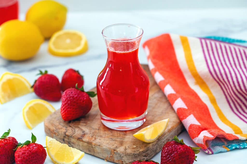 Head-on view of a glass bottle of strawberry lemonade syrup with lemons and strawberries all around.
