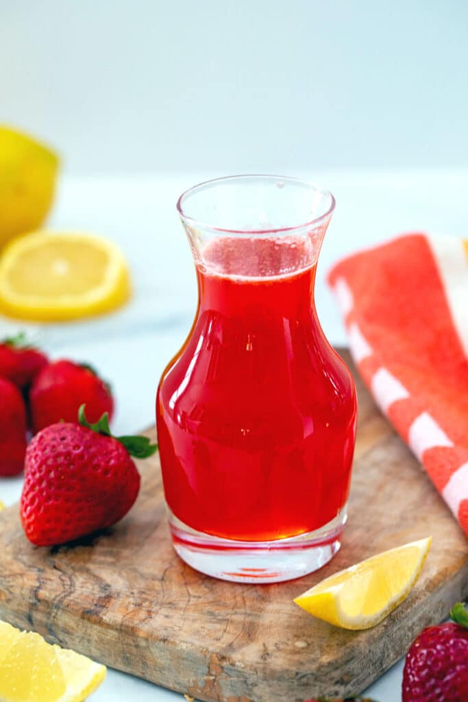 Head-on view of a small glass filled with red strawberry lemonade syrup with lemon wedges and strawberries surrounding it.