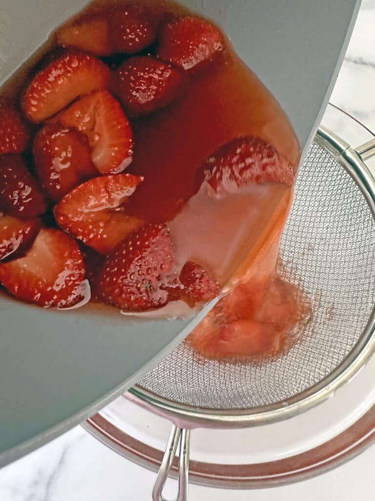 Strawberry syrup being poured from saucepan into strainer.