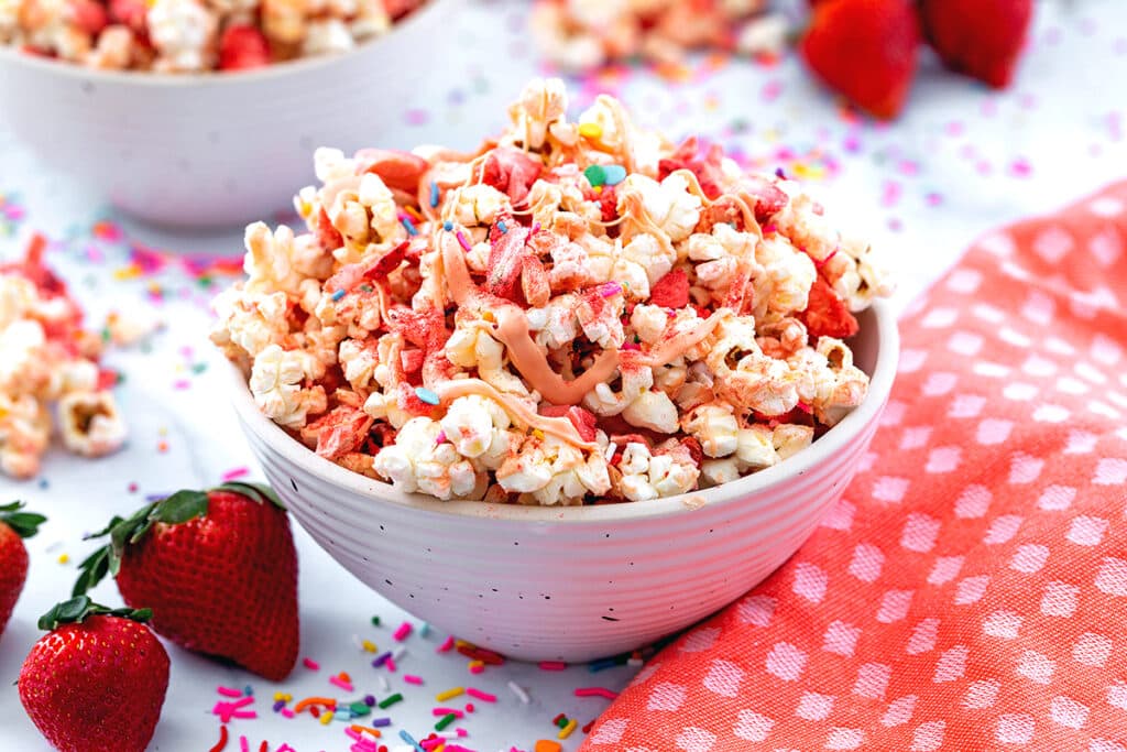 Landscape head-on view of bowl of strawberry popcorn with fresh strawberries and sprinkles all around.