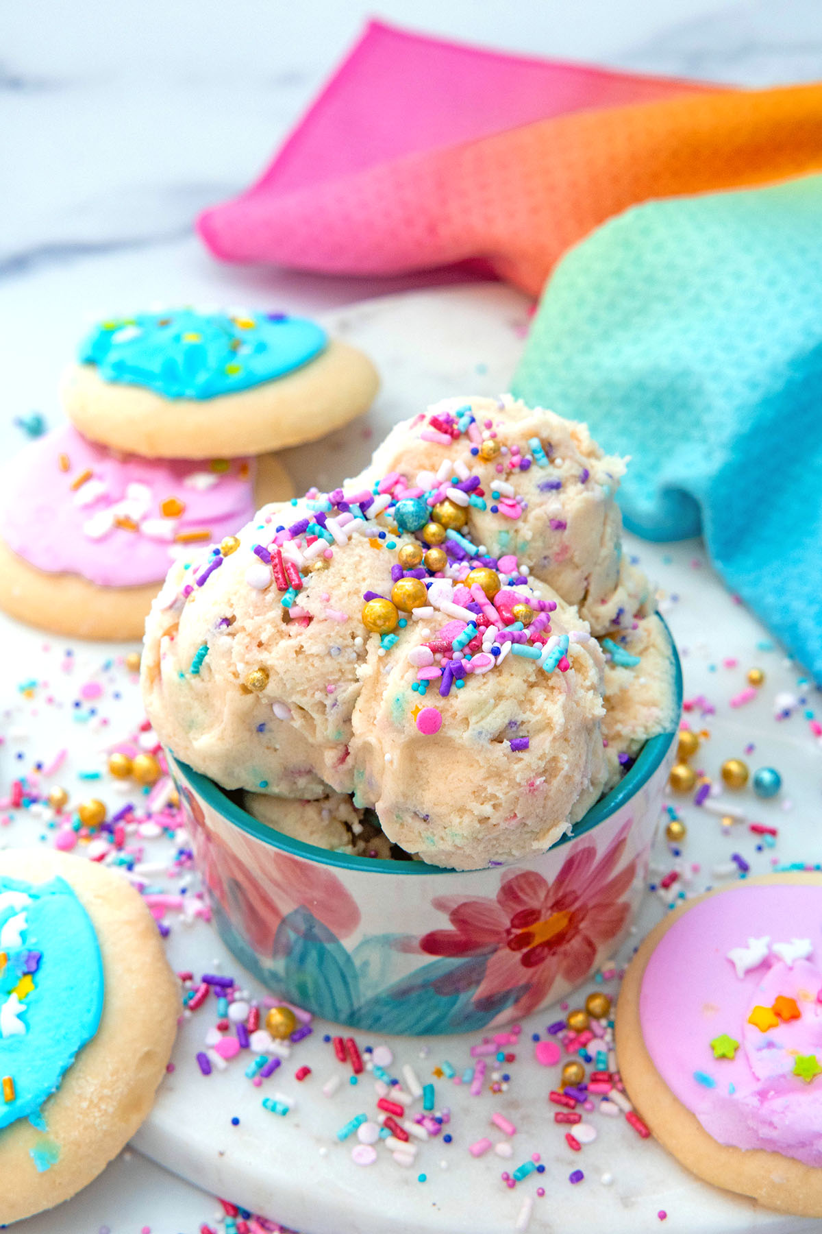 Overhead view of a bowl of sugar cookie dough with sugar cookies and pastel sprinkles all around.
